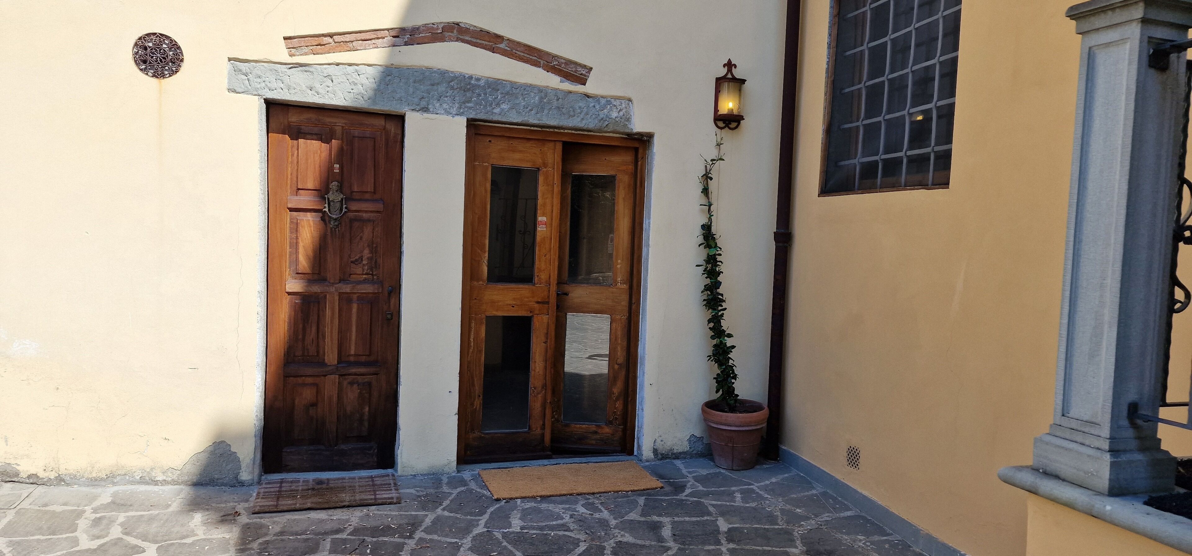 A stone-paved entrance with two wooden doors, a potted plant, and a wall-mounted light fixture.