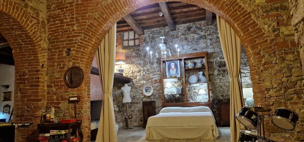 A rustic room with exposed brick walls, a wooden ceiling, a bed with a white bedspread, a dresser with bottles, a lamp, a mannequin, and a shelf with decorative items.