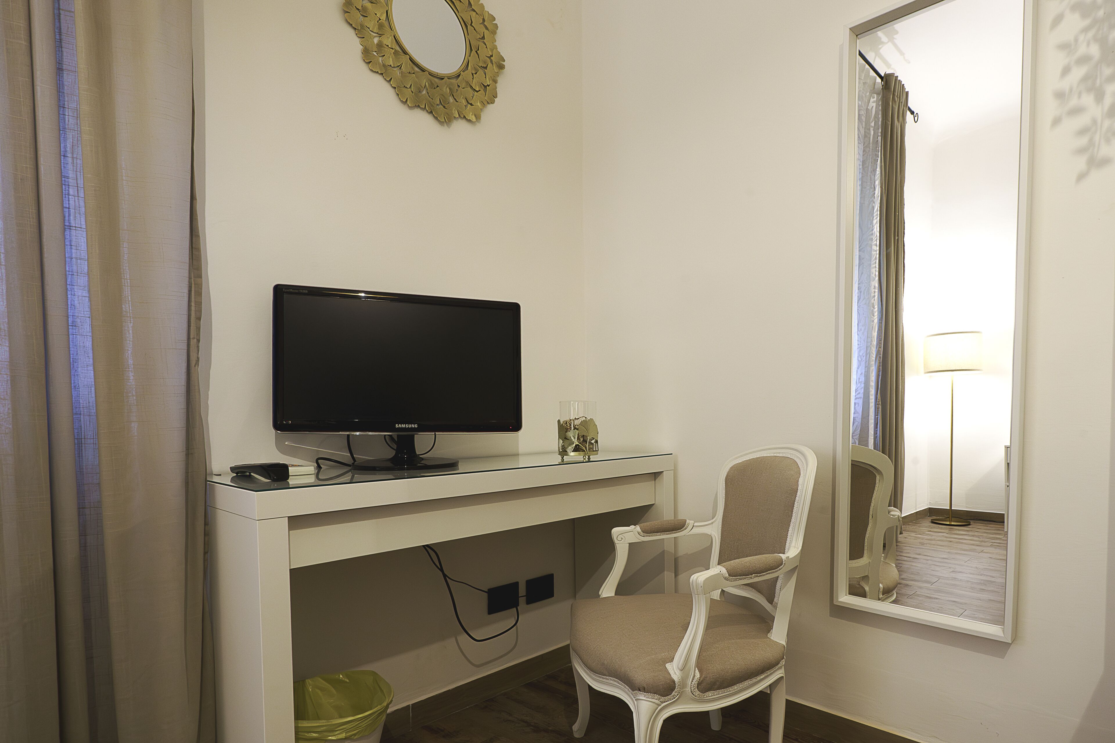 Een kamer met een wit bureau, een televisie, een stoel, een prullenbak en een spiegel.