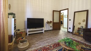 Ein Wohnzimmer mit einem Flachbildfernseher auf einem weißen Standfuß, Holzboden, einem gemusterten Teppich und einer Tür zu einem anderen Raum.