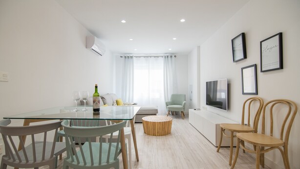 Uma sala de estar moderna com uma mesa de jantar de vidro, cadeiras de madeira, um banquinho de madeira redondo, um sofá e uma TV fixada na parede.