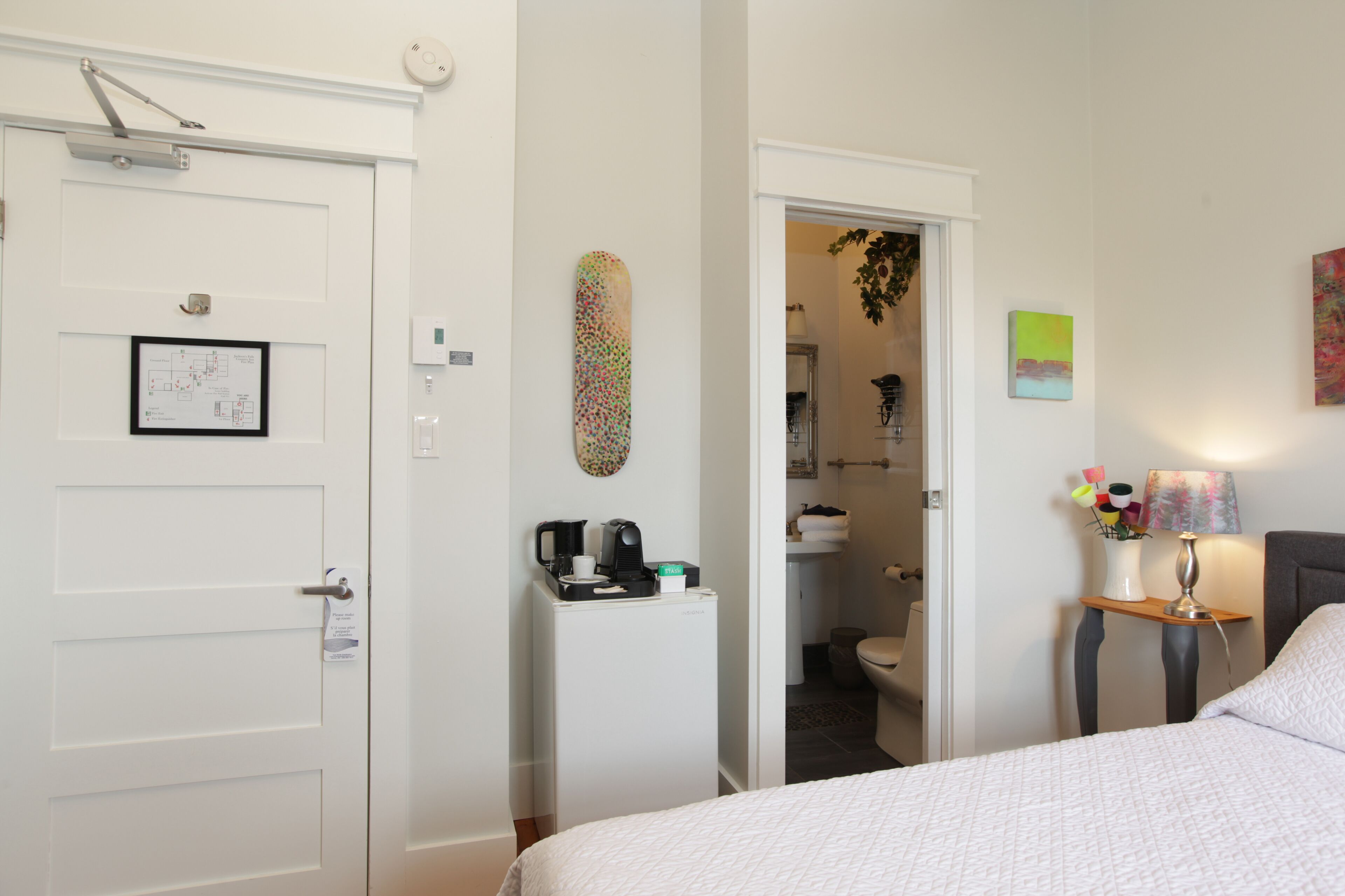 Hotelkamer met een bed, een bureau met lamp, een koelkast en een deur met een fotolijstje en een bordje.