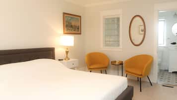 A hotel room with a bed, two chairs, a nightstand with a lamp, a painting, a window with a view, and a mirror.