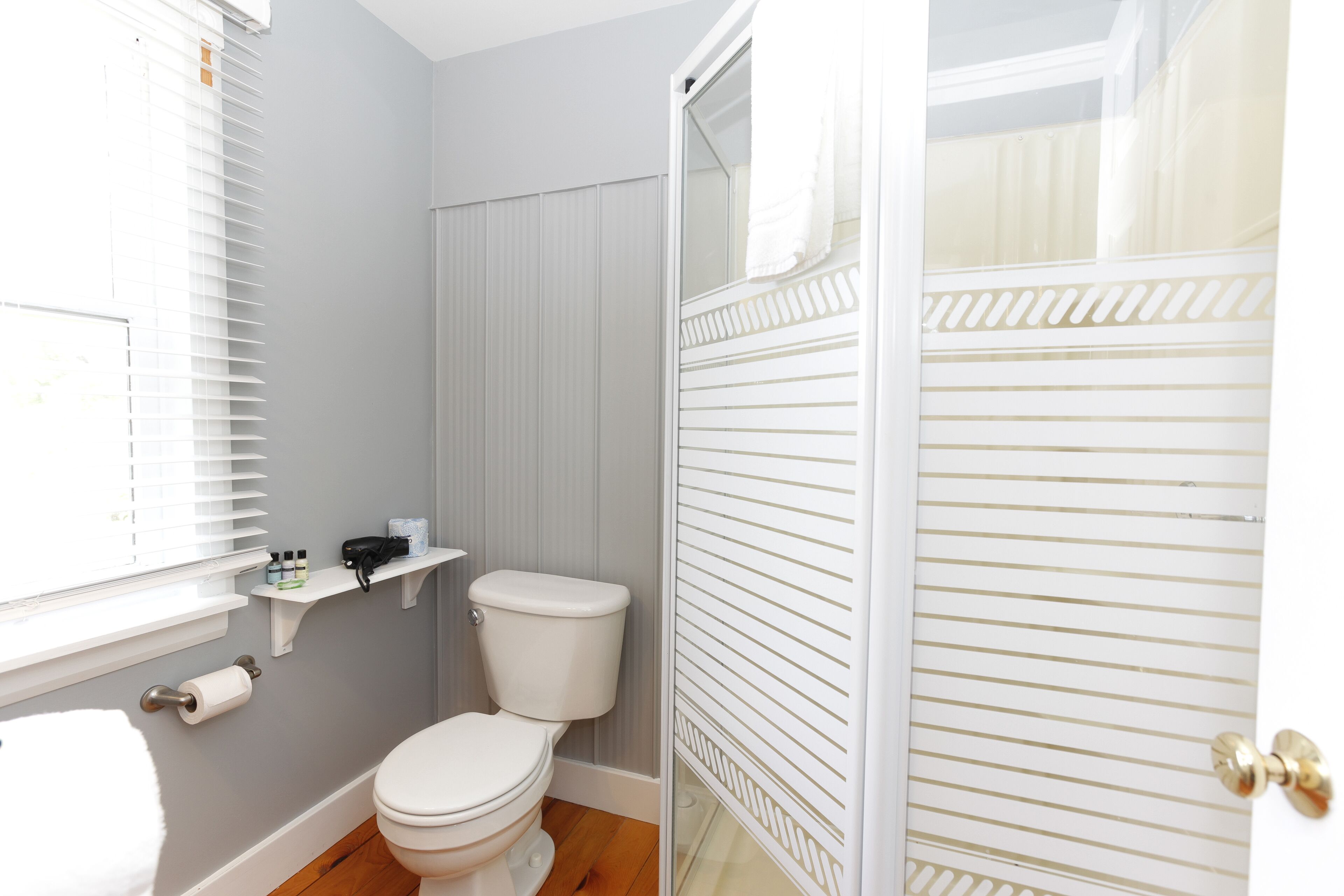Un baño con inodoro, ducha, ventana con persianas y un perchero para toallas.