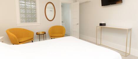 A bedroom with a bed, two yellow chairs, a small round table, a TV mounted on the wall, and a door leading to a bathroom.