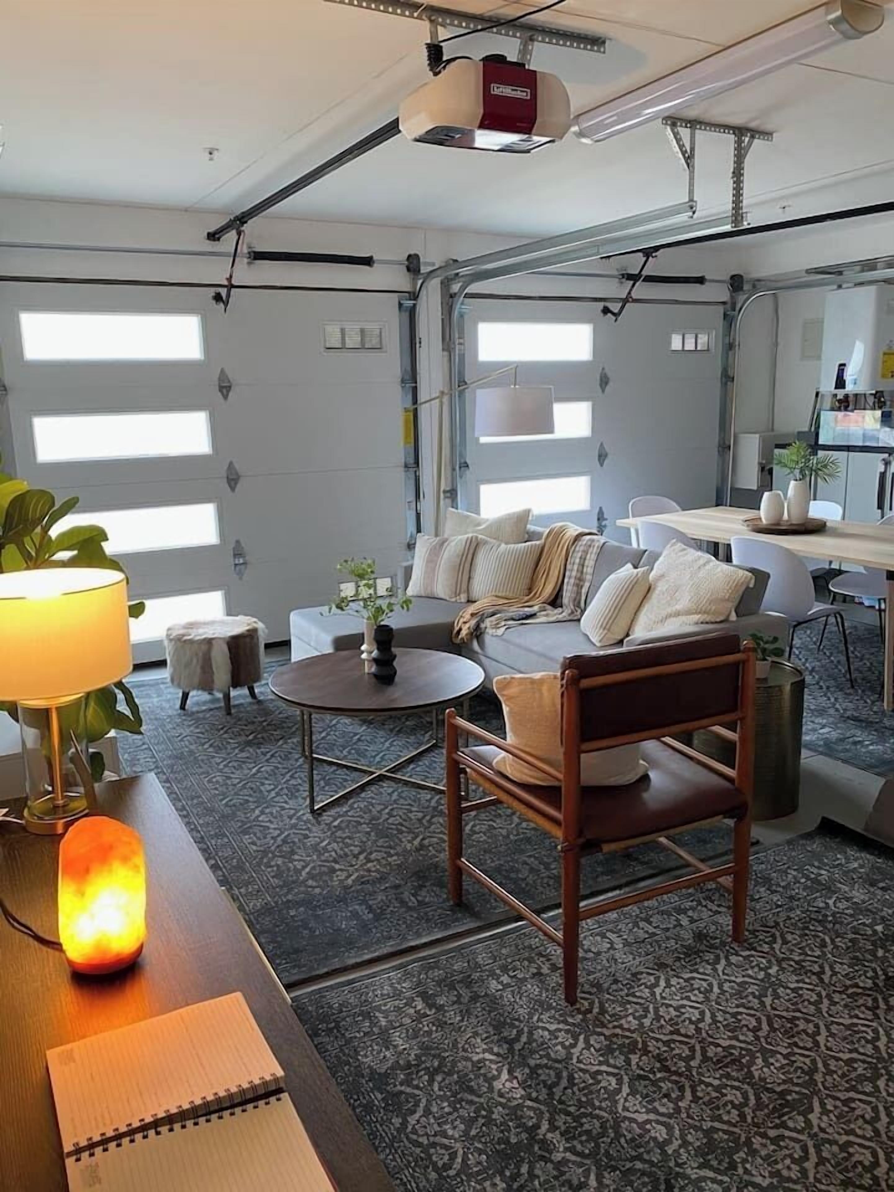 Een moderne, goed verlichte woonkamer met een bank, salontafel en een lamp. Er staat een klein rond tafeltje met een plant en een notitieboekje. De kamer heeft grote ramen en een zichtbare keukenhoek.