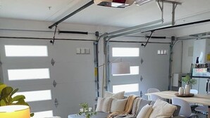 A modern, well-lit living room with a sofa, coffee table, and a lamp. There is a small round table with a plant and a notebook. The room has large windows and a visible kitchen area. - Luxury 7BR Home Near SFO w/Parking (Daly City)