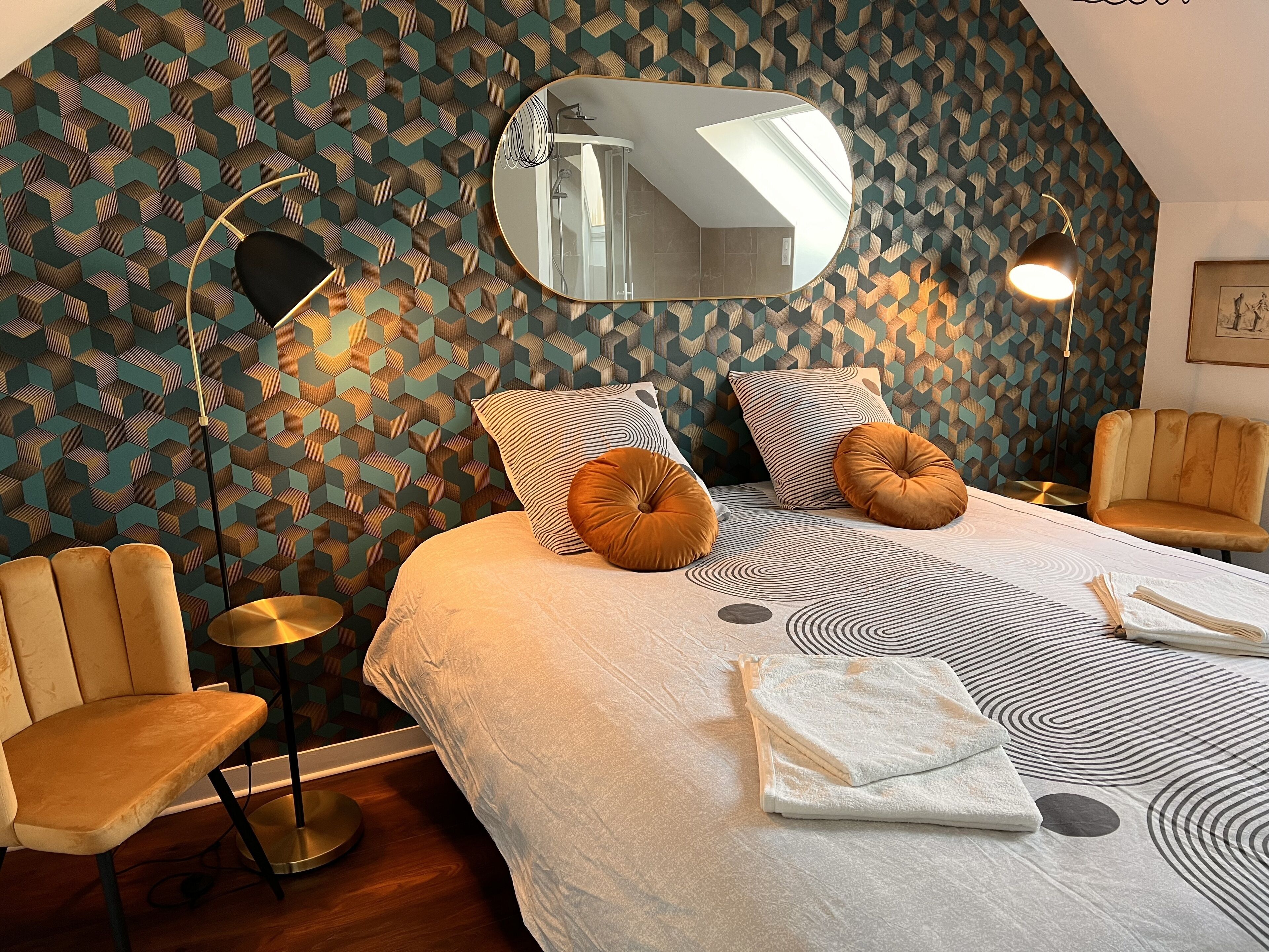 Une chambre moderne avec un mur orné d’un motif géométrique, un grand lit avec une literie à motifs, deux fauteuils et une lampe sur pied.