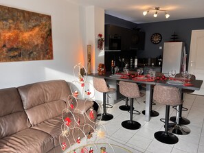 A modern living room with a brown leather sofa, a dining area with a long table set for six, and a kitchen in the background. - Cosy house near CO'Met (Orléans)