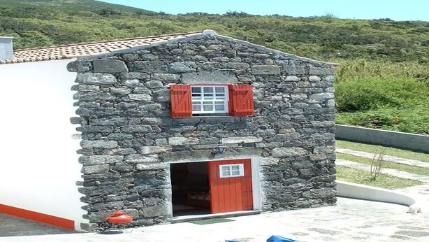 Ein Steinhaus mit roter Tür und Fensterläden, umgeben von viel Grün.