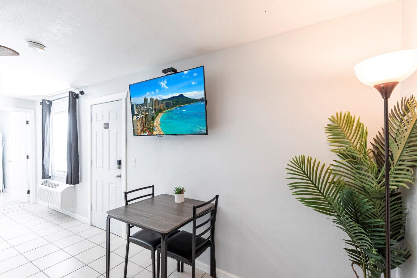 Habitación de hotel con comedor que incluye una mesa y sillas, un televisor montado mostrando una vista panorámica, una planta en maceta y una lámpara de pie.