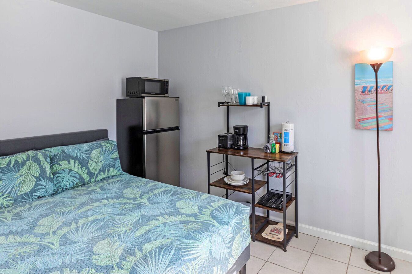 A hotel room with a bed, a refrigerator, a coffee maker, a shelf with glasses and a coffee pot, and a painting on the wall.