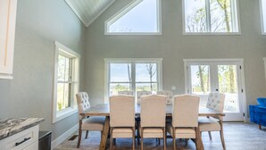 A dining area with a wooden table and six upholstered chairs, large windows, and a view of trees. - Luxury Mountain Escape (Hendersonville)