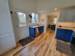 Una cocina con suelo de madera, armarios azules, un fregadero y un comedor visible a través de una puerta abierta.