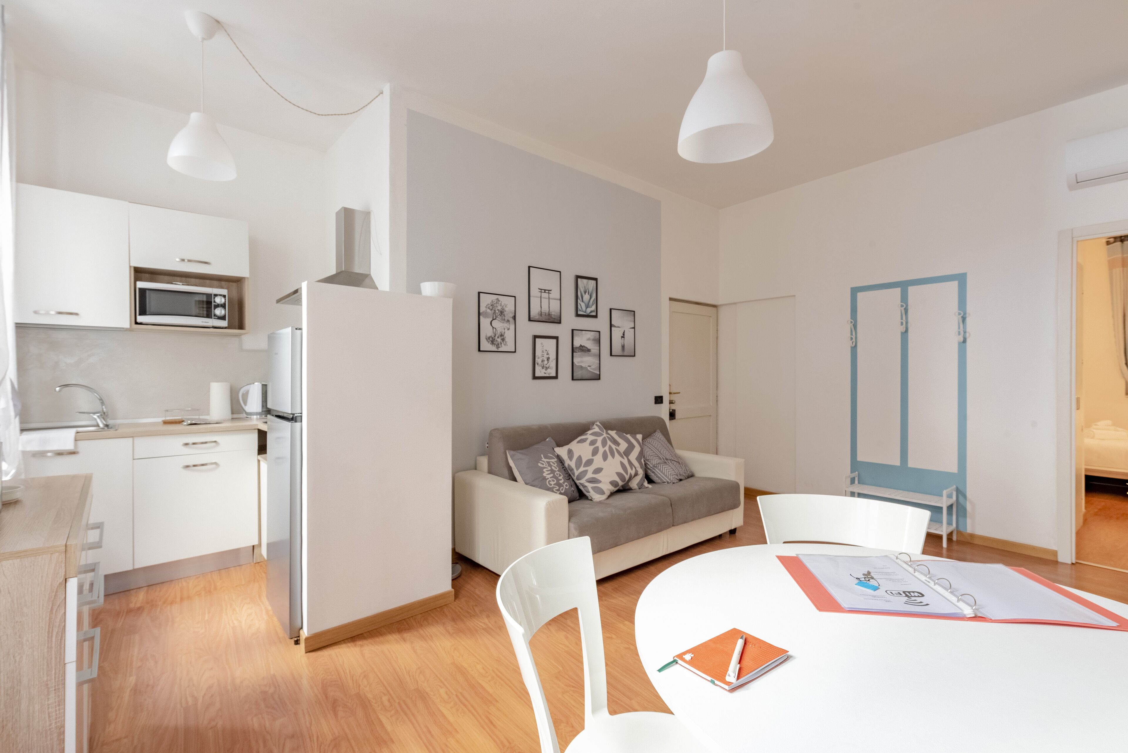 Una cocina moderna con armarios blancos, un microondas y un refrigerador. Hay una zona de comedor con una mesa y sillas, y un sofá al fondo.