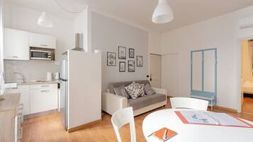 Una cocina moderna con armarios blancos, un microondas y un refrigerador. Hay una zona de comedor con una mesa y sillas, y un sofá al fondo.