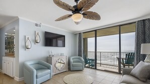 A spacious living room with a ceiling fan, a flat-screen TV, a round coffee table, and a balcony with a view. - The Whaler D5 by Liquid Life (Gulf Shores)