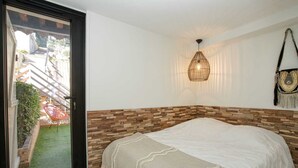 Een slaapkamer met een houten hoofdbord, een bed met een witte dekbedovertrek, een geweven lamp en uitzicht op een tuin door een schuifdeur.