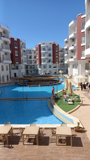 A swimming pool area with lounge chairs and umbrellas, surrounded by multi-story buildings with balconies. - Aqua palms resort hurghada  (Hurghada)
