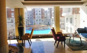 A modern living room with a large window overlooking a swimming pool and residential buildings. - Aqua palms resort hurghada  (Hurghada)