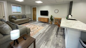 A modern living room with a sofa, a coffee table, a TV, a kitchen area with a sink, and a dining table. - 2 bedroom Budget Suite. Open floor plan (Mattoon)