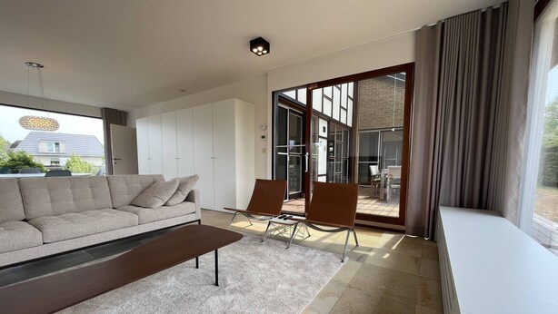 Un salon moderne avec un canapé, une table basse et deux fauteuils. Une porte-fenêtre coulissante donne accès à un espace extérieur.