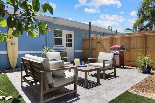 A patio with two beige cushioned chairs and a small table, a wooden fence, and a barbecue grill.