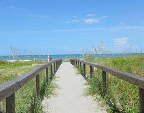 En träpromenad som leder till en strand med högt gräs och en klar, blå himmel.