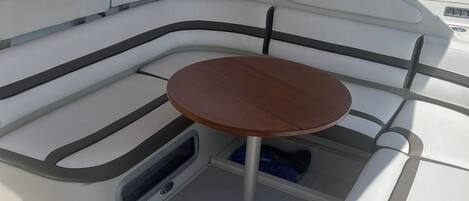 Interior de un barco con una zona de estar acolchada, una mesita pequeña y un compartimento para guardar objetos.