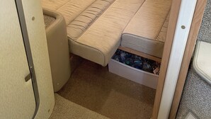 A compact, beige-colored sleeping area with a bench-style bed, a storage compartment below, and a ceiling vent.