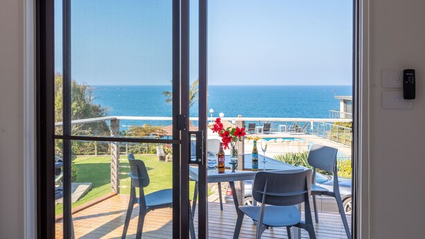 A balcony with a view of the sea, a table with chairs, and a bottle of beer. - Breathtaking Clifftop Ocean Views in Eden - Unit 12 Bob and Zita’s escape (Eden)