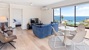 A modern living room with a blue sofa, a TV, and a dining area with chairs and a table. - Breathtaking Clifftop Ocean Views in Eden - Unit 12 Bob and Zita’s escape (Eden)