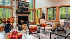 A cozy living room with a stone fireplace, comfortable seating, and a large window overlooking a forest. - North GA Relaxing Chic Cabin w/ Resort Amenities (Ellijay)