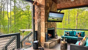 A stone fireplace with a TV, outdoor seating, and a hot tub on a wooden deck. - North GA Relaxing Chic Cabin w/ Resort Amenities (Ellijay)