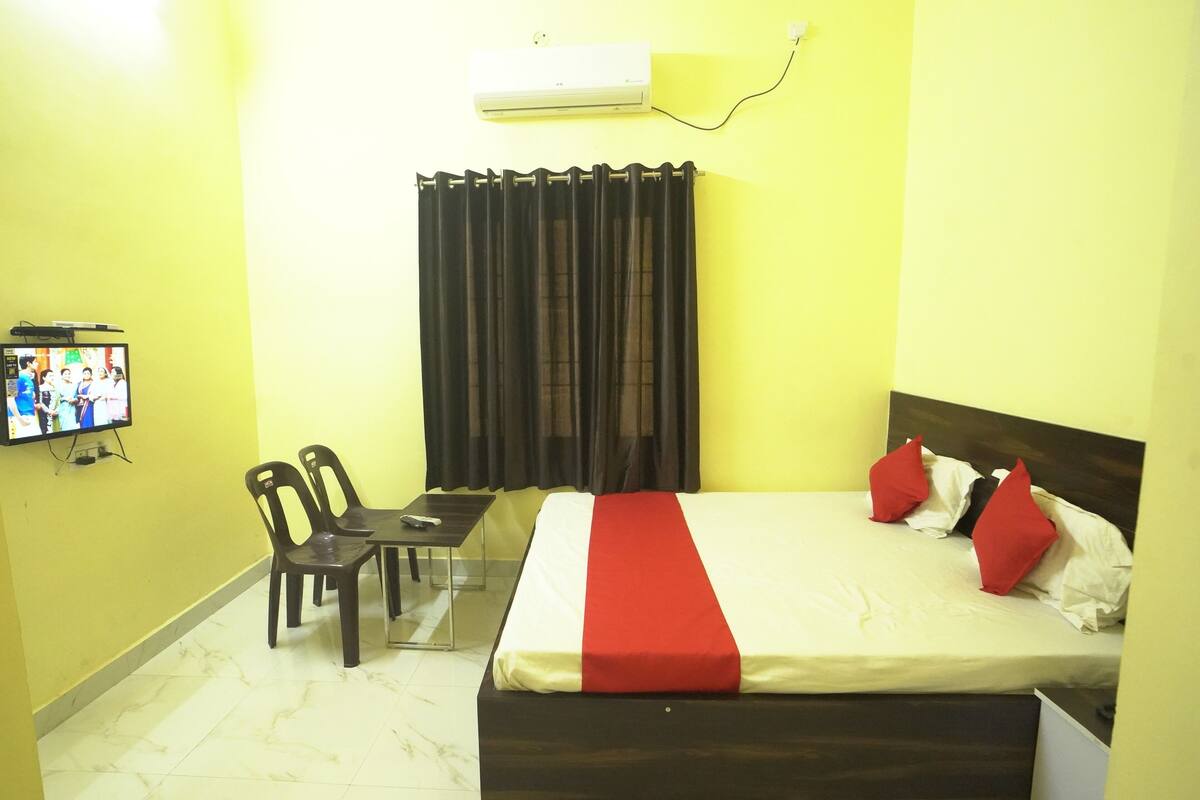 Una habitación de hotel con cama, televisión sobre un mueble, una mesa con sillas y una cortina.