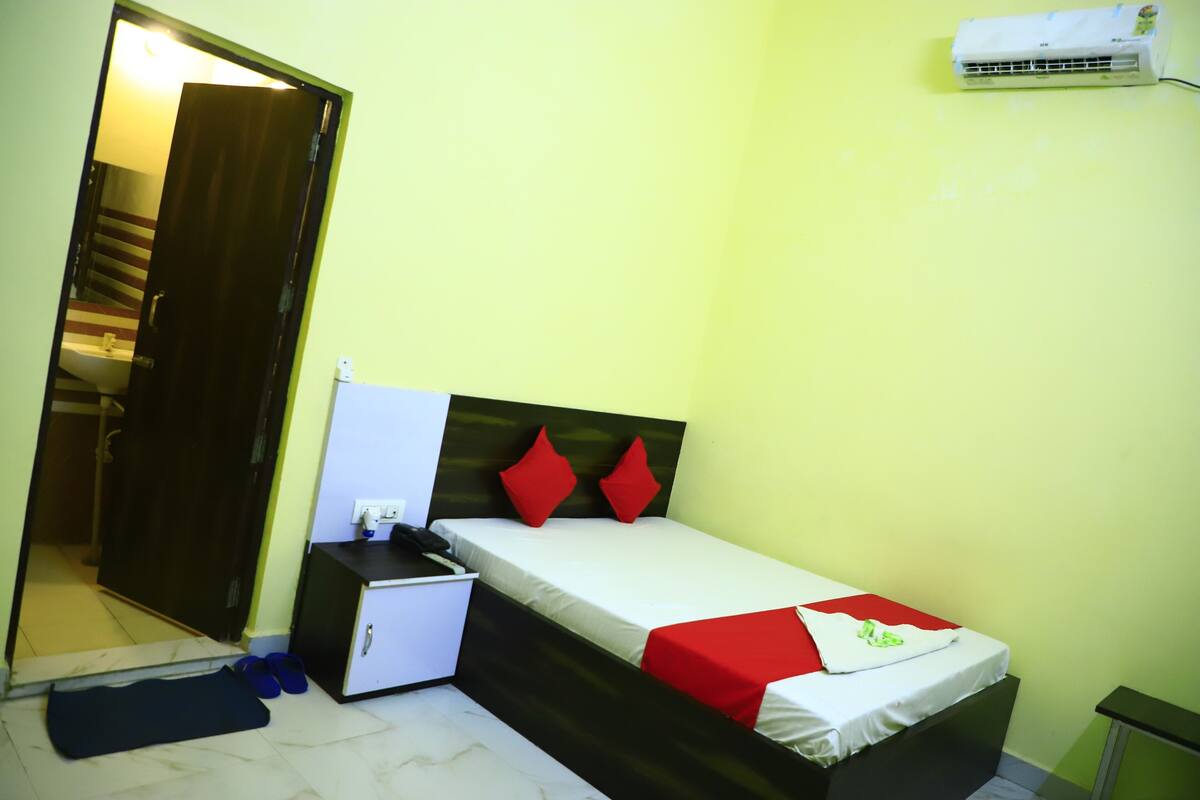 Una habitación pequeña con cama individual, colchón blanco, almohadas rojas, un escritorio con silla, un aire acondicionado blanco y una puerta que da a un baño.