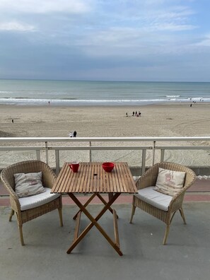 Un patio frente a la playa con sillas de mimbre y una mesa de madera, con vistas a una playa arenosa donde se ven personas a lo lejos.