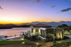 A modern house with a stone exterior, a glass balcony, and a view of the sea at sunset. - Panoramic Sea Views to Spinalong Island with a heated pool (Elounda)