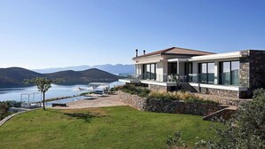 Een modern huis met een stenen gevel, grote glasramen en uitzicht op een meer en bergen.