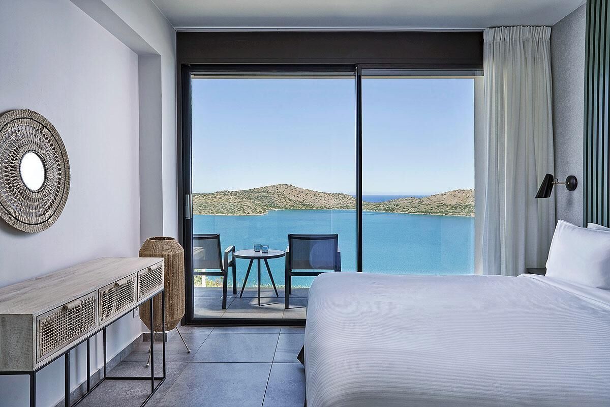 Quarto de hotel com uma cama grande, uma escrivaninha com cadeira, uma mesinha com duas cadeiras e vista para o mar e as colinas.