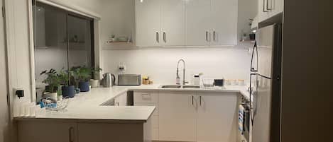 Cocina moderna con armarios blancos, encimera de mármol, lámpara de araña y refrigerador empotrado.