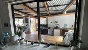 A modern kitchen with a built-in grill, a dining area with a wooden table, and a view through a glass door to an outdoor space. - Newly Renovated Hamptons-Style Beach House - Includes Linen & EV Charger! (Coronet Bay)