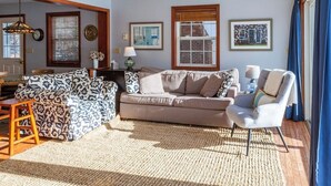 A living room with a patterned sofa, a neutral-toned sofa, and a chair. There are framed pictures on the wall, a window with blinds, and a wooden floor.