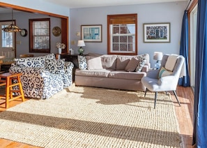 A living room with a patterned sofa, a neutral-toned sofa, and a chair. There are framed pictures on the wall, a window with blinds, and a wooden floor. - Chickadee Bay House- Oceanfront! (Trenton)