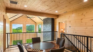 A wooden cabin with a hexagonal table and chairs, a balcony with a view of the forest, and a stone fireplace. - Spacious & Upscale with 7 Person Hot Tub, Theater, Views, Game Room, Sauna (Sevierville)