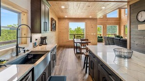 A modern kitchen with wooden beams, a dining area with a view, and a cozy seating area with a clock on the wall. - Spacious & Upscale with 7 Person Hot Tub, Theater, Views, Game Room, Sauna (Sevierville)