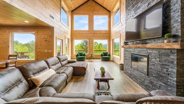 A spacious living room with a leather sectional sofa, a fireplace, and large windows offering a view of the outdoors. - Spacious & Upscale with 7 Person Hot Tub, Theater, Views, Game Room, Sauna (Sevierville)