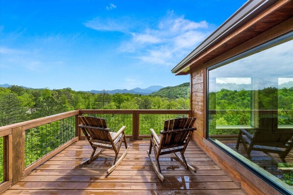 A wooden deck with rocking chairs overlooking a lush green landscape. - Spacious & Upscale with 7 Person Hot Tub, Theater, Views, Game Room, Sauna (Sevierville)