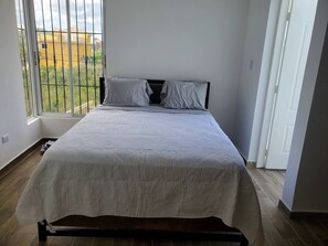 A single bed with a white bedspread in a room with a large window, wooden flooring, and a view of buildings outside.