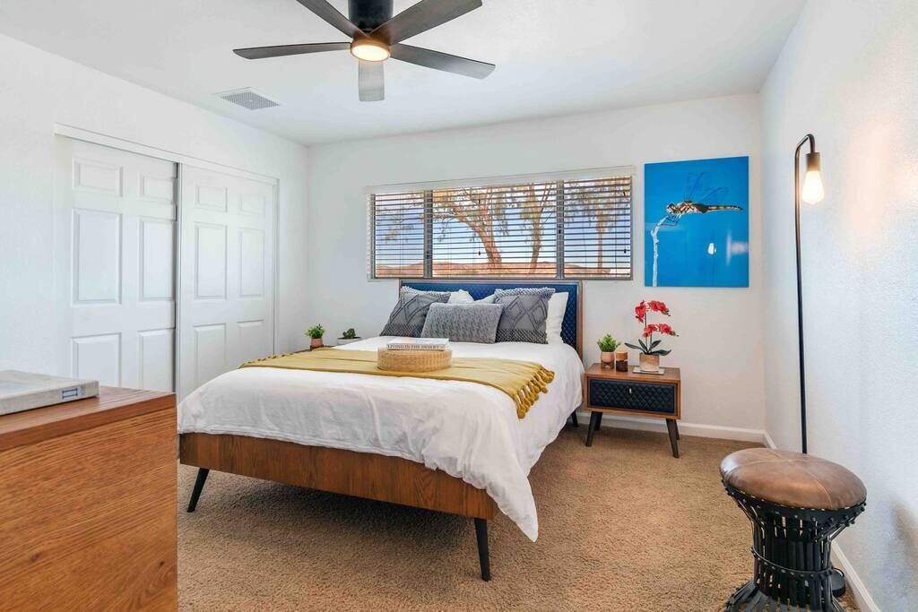 Une chambre à coucher comprenant un lit, un ventilateur de plafond, une table de chevet, un banc, une lampe fixée au mur et une fenêtre avec des stores.